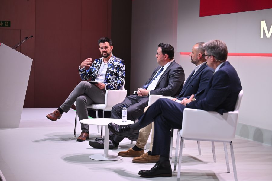 Four men in suits sit on stage having a panel discussion, with one man speaking and gesturing with his hand.