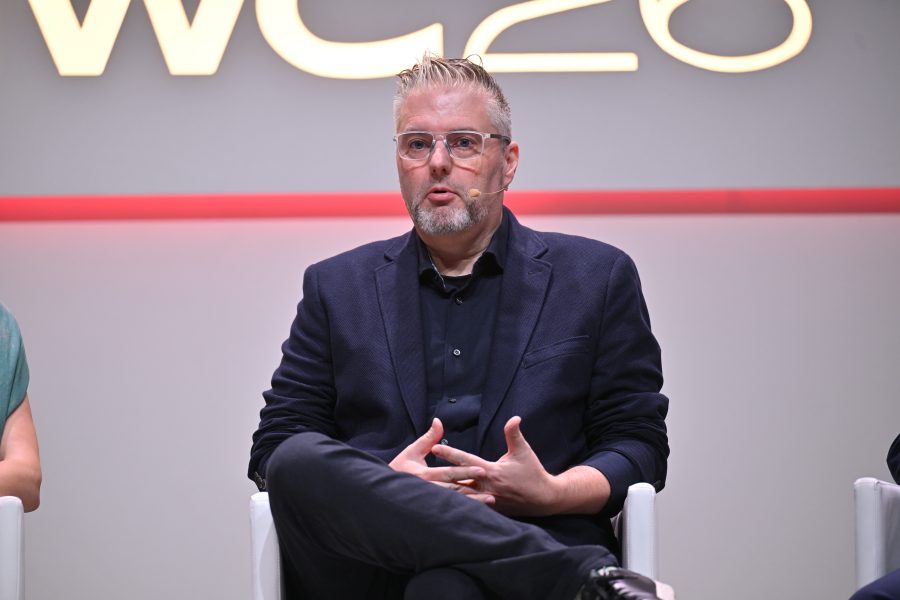 A man with glasses and a headset speaks on stage, seated with hands clasped, against a lit-up backdrop.