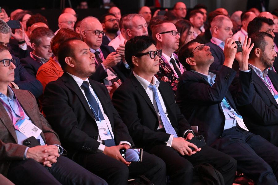 A group of people in business attire sit closely together at a conference, some holding mobiles or taking photos.