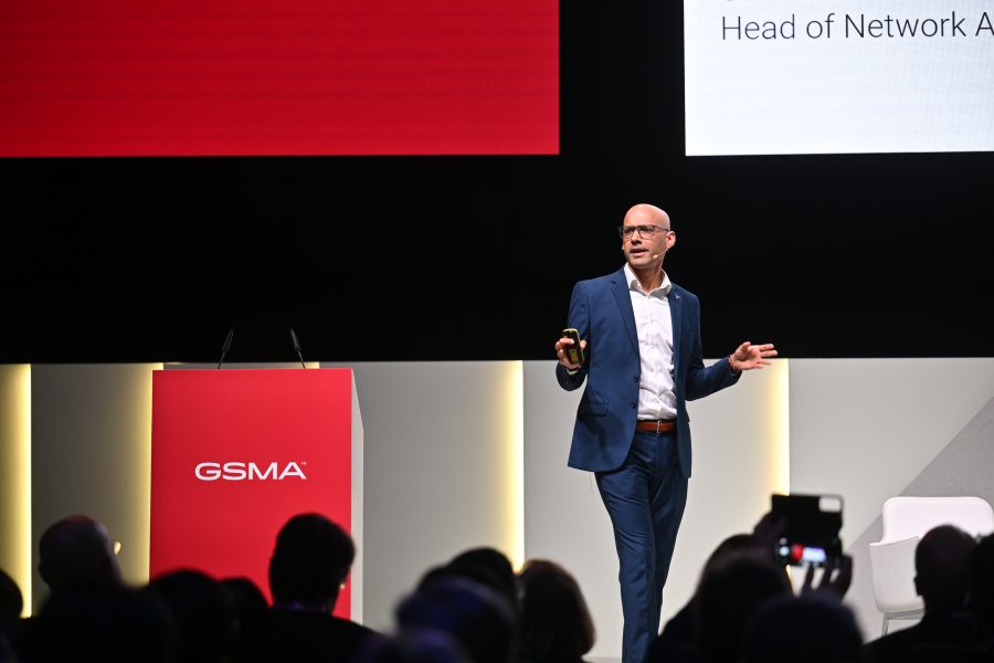 Bald man in a blue suit speaks on stage at a conference, with a red GSMA lectern beside him.