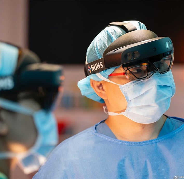 A surgeon wearing a face mask, blue surgical gown, cap, and augmented reality headset labelled "NUHS" focuses intently. Another masked medical professional is visible in the blurred background. The scene suggests a high-tech medical environment.