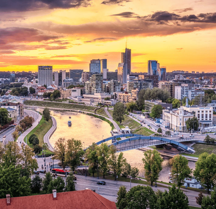 A vibrant cityscape at sunset shows modern high-rise buildings and older structures along a winding river. A pedestrian bridge crosses the water, and lush trees line the riverbanks. The sky glows orange and yellow, casting warm light over the urban scene.