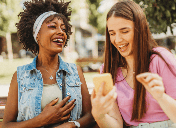 Two young women sit on a bench outdoors, laughing together as they look at a mobile phone. One has curly hair with a headband and wears a denim waistcoat, while the other has straight brown hair and a pink top. Trees and blurred buildings appear in the background.