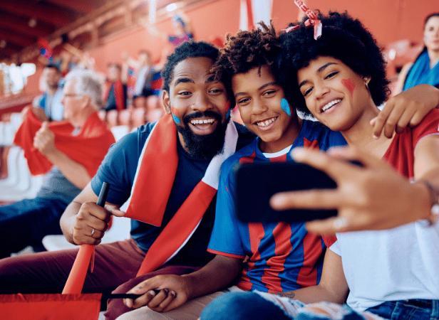 A smiling family of three takes a selfie at a stadium. They wear face paint and team colours, celebrating with flags. The stands are filled with supporters, and the atmosphere appears joyful and energetic.