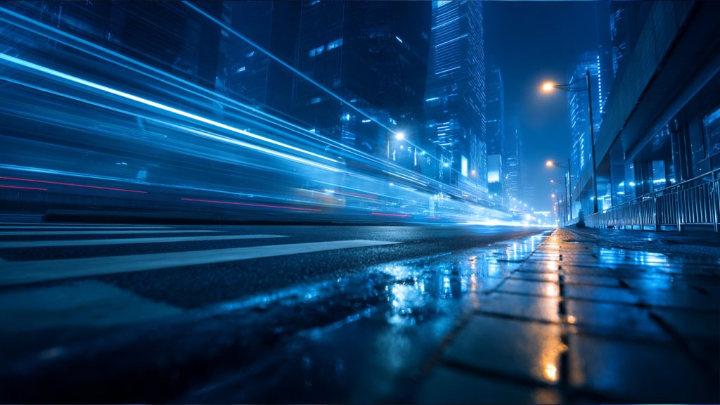 A wet city street at night pulses with bright blue and white light trails from fast-moving vehicles. Tall glass buildings reflect neon lights, creating a futuristic urban scene that hints at the impact of quantum technologies on modern life.