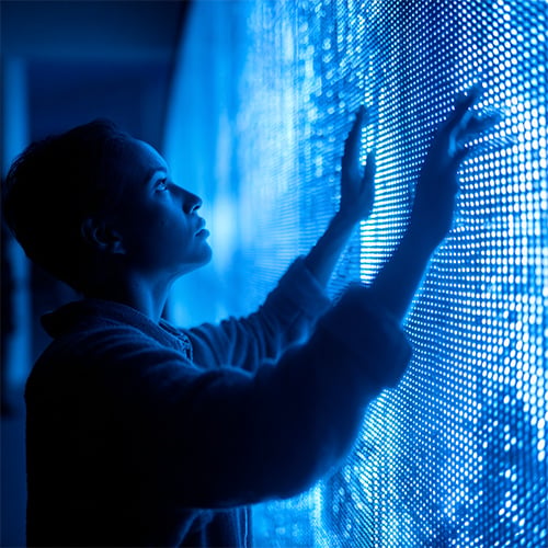 A person stands in profile, touching a large illuminated wall of tiny blue lights. Blue light bathes the scene, casting a futuristic glow as they thoughtfully engage with the glowing, digital-looking surface inspired by quantum technologies.