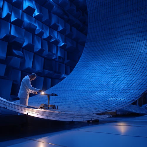 A person in a white lab coat examines equipment in a large, futuristic blue room with curved, tiled walls and geometric acoustic panels—a dramatic scene highlighting the high-tech, scientific world of quantum technologies.