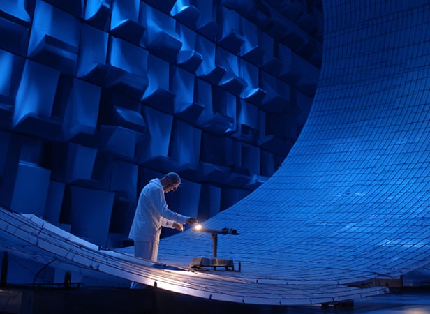 A person in a white lab coat testing quantum communication under a bright light in a large, futuristic blue chamber with geometric, sound-absorbing panels and curved metallic surfaces, suggesting a high-tech laboratory or testing facility.