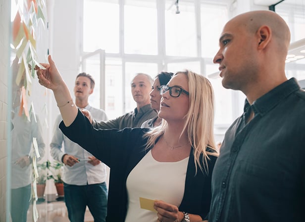 Five people from the Quantum Network Service Working Group stand in a bright office, discussing and placing sticky notes on a glass wall. A woman in glasses points to a note whilst another man looks on closely. Three others observe the wall, engaged in the collaborative activity.