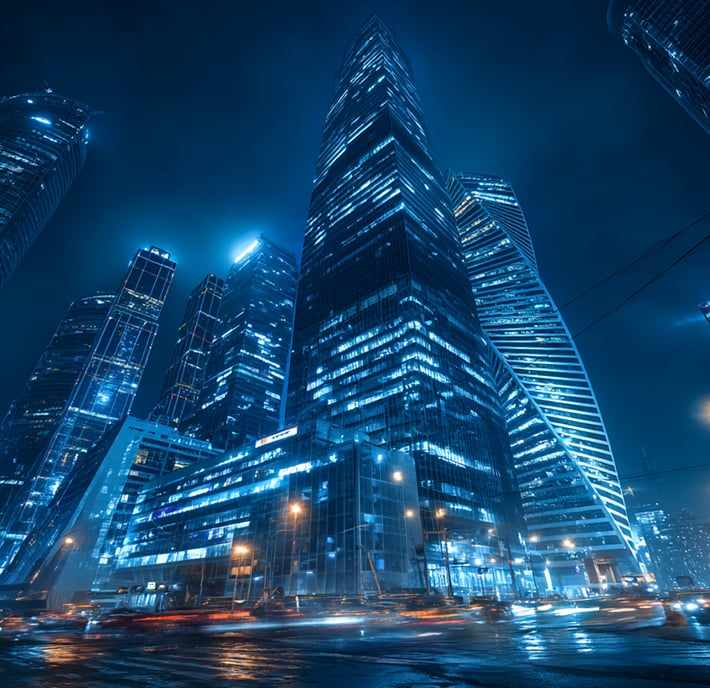 A night-time cityscape with modern skyscrapers illuminated by blue lights. The tall glass buildings reflect the glow, whilst blurred car lights show quantum communication