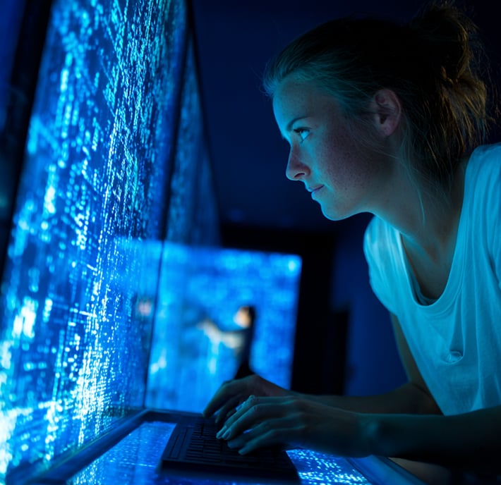 A woman in a white shirt leans over a keyboard, focused on quantum communication screens displaying complex digital code and data, creating a high-tech, futuristic atmosphere in a dark room.