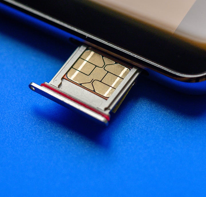 A close-up of a SIM card tray partially ejected from a mobile phone on a blue surface. The gold contacts of the SIM card are clearly visible, and part of the phone’s edge and screen can be seen in the upper right corner.