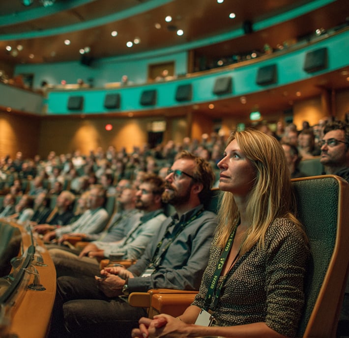 A large audience sits attentively in a modern, dimly lit auditorium in a scam prevention seminar. The focus is on a woman in the foreground, watching the stage intently. People of various ages are seated around her, all looking forward. The atmosphere is serious and engaged.