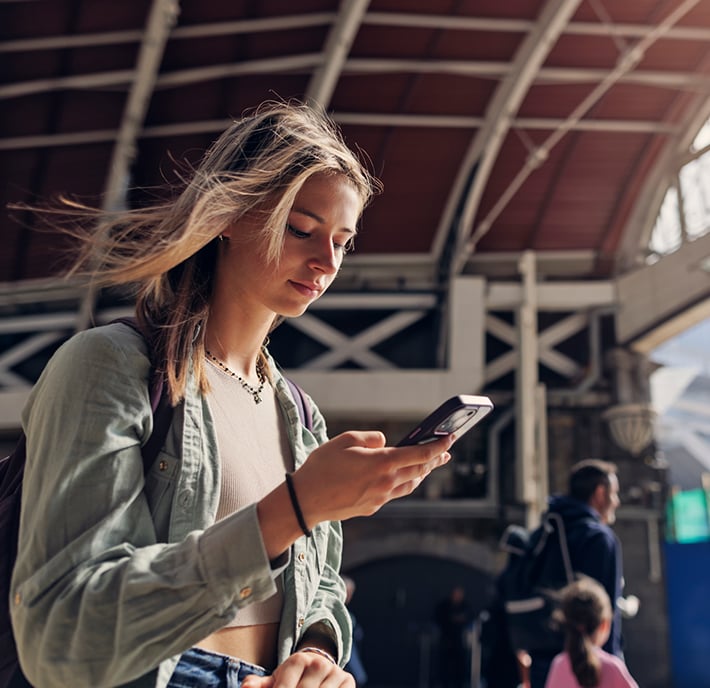 A young woman with light brown hair stands in a railway station, looking at her mobile phone. She wears a light green shirt and a necklace, and carries a rucksack. The station has a curved roof and other people are blurred in the background.