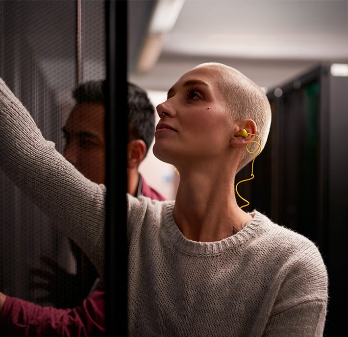 A person with a shaved head, wearing a light jumper and yellow earplugs, reaches into a server rack in a dimly lit data centre. Another person in the background is partly visible, focused on the equipment.