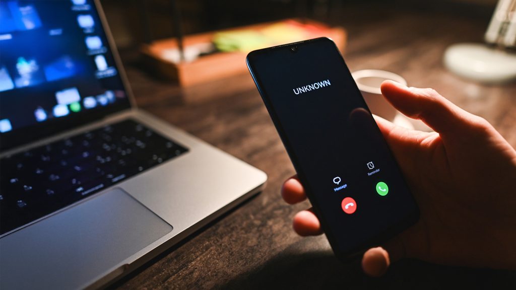A hand holds a mobile phone showing an incoming call from "UNKNOWN" with green and red buttons for answering or rejecting—highlighting scam prevention. The phone rests by an open laptop on a dimly lit wooden desk, white mug nearby.