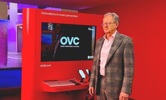 Man in a checked suit stands beside a red exhibition stand with "OVC" and "Innovations in scam prevention" on the screen.