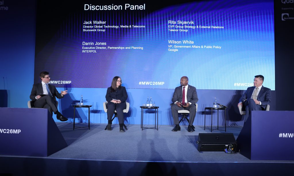 Four panellists sit on stage in discussion, with their names and titles displayed on a screen behind them.