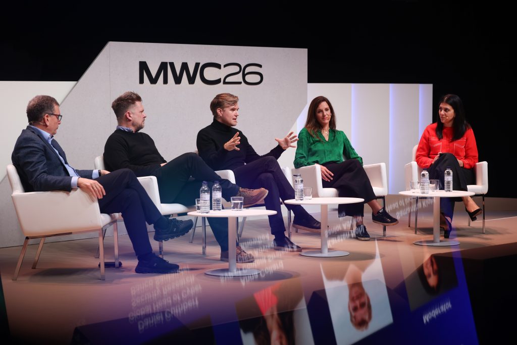 Five people sit on stage in discussion at the MWC26 event, with water bottles on tables in front of them.