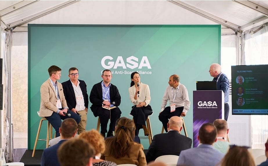 Five people sit on stools on a stage in front of a green “GASA” backdrop; one woman speaks into a microphone. An audience watches, and a man stands at a lectern with a screen beside him inside a marquee event space.