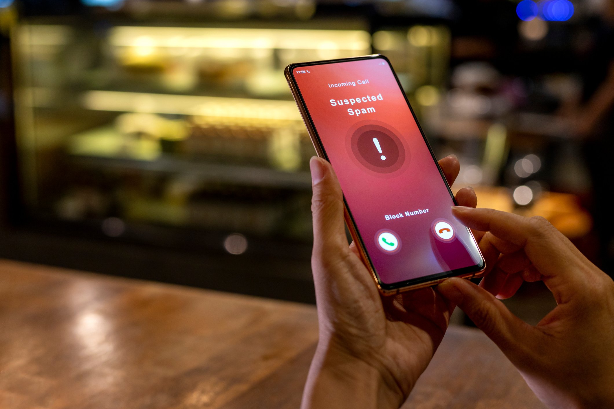 A person holds a mobile phone displaying an incoming call screen that reads “Suspected Spam,” with large exclamation mark icon, and options to accept or reject the call. The phone is held over a wooden table with blurred indoor café background.
