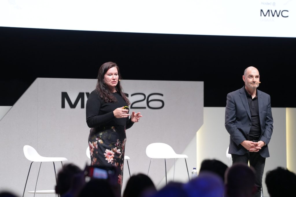 A woman speaks on stage next to a man at MWC 2026, with two empty chairs behind them.