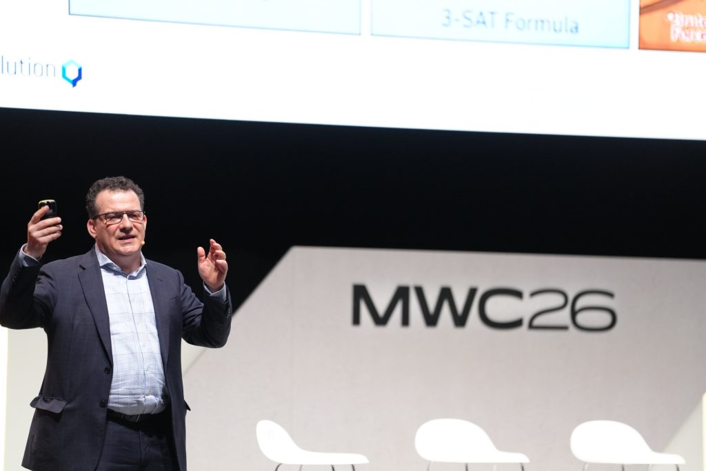 A man in a suit speaks on stage at MWC26, with three empty chairs in the background.