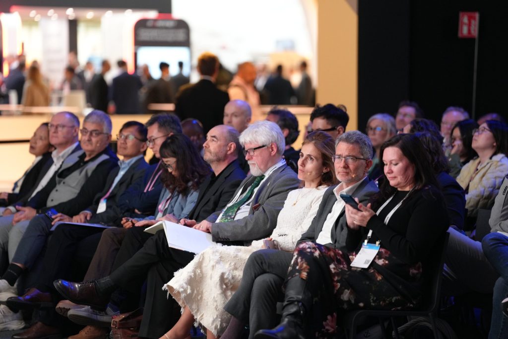 Audience members sit attentively in rows at a conference, some taking notes or using their mobiles.