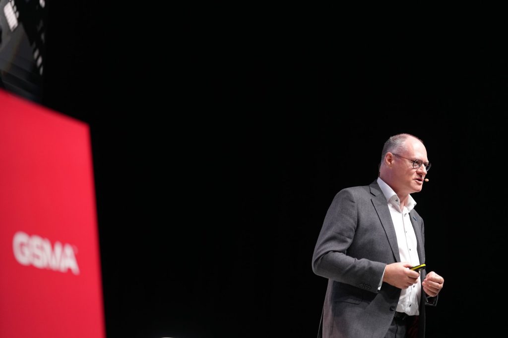 A man in a suit speaks on stage with a microphone; a red sign with "GSMA" is visible on the left.