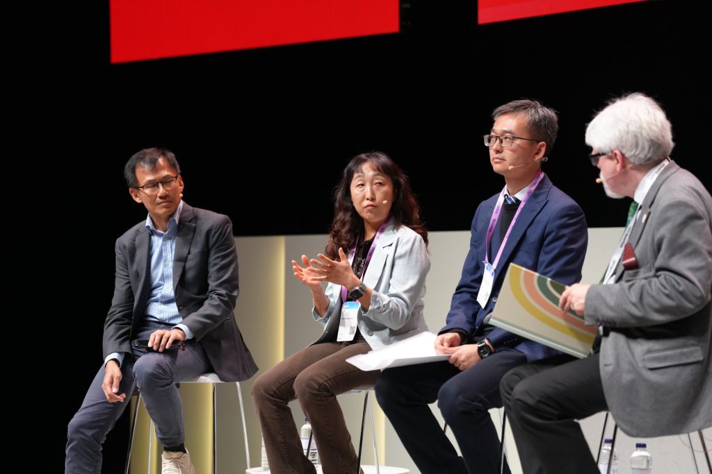 Four people sit on stools and engage in a panel discussion on stage with papers and microphones.