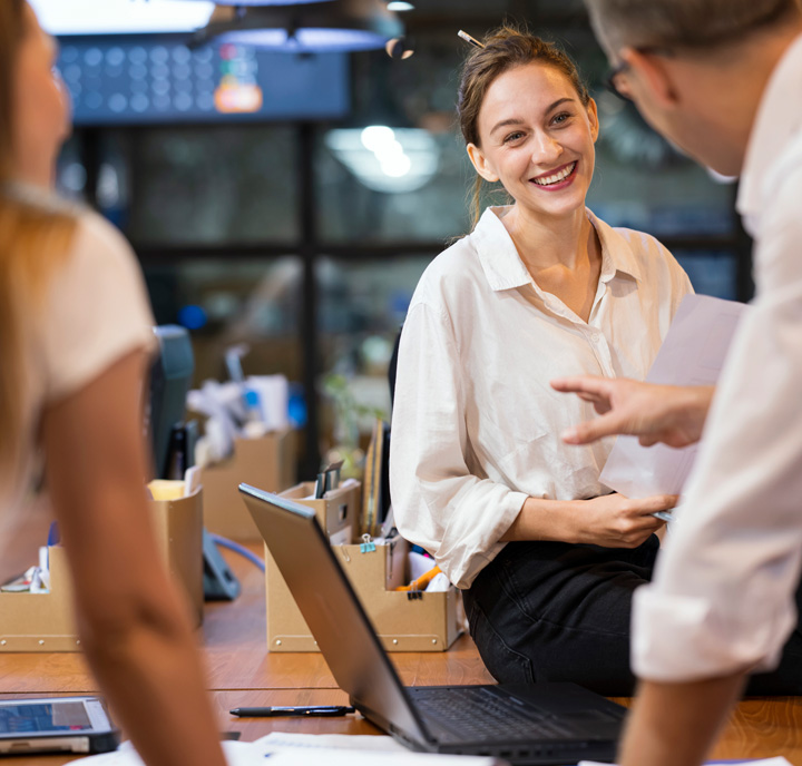 Two colleagues engage in a lively conversation with a smiling woman sitting on a desk. She holds papers, dressed in a white blouse and black pants. Laptops and office supplies are scattered around. The atmosphere is light and friendly, indicating a collaborative work environment.