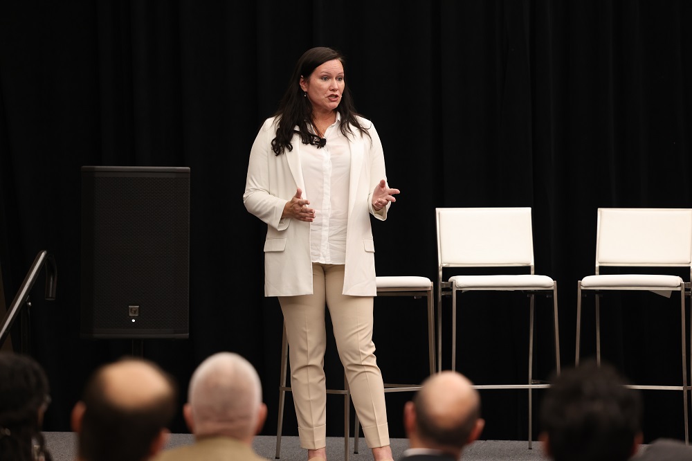A woman in a white blazer and beige pants speaks on a stage with a black backdrop. She is gesturing with her hands. Three white chairs and a large speaker are visible behind her. The heads of several audience members are in the foreground.