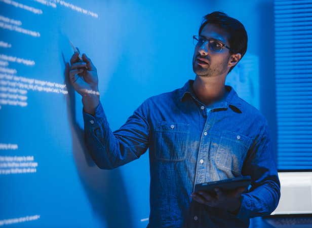 A man in glasses and a denim shirt stands in front of a blue-lit wall covered with code or text. He holds a tablet in one hand and points to the wall with the other, suggesting engagement in analysis or teaching. The setting appears to be a modern, tech-oriented work environment.