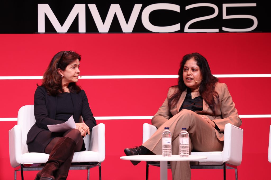 Two women are seated on white chairs, engaged in a conversation about post-quantum security in front of a red backdrop displaying "MWC25". The woman on the left has papers and wears a dark outfit. The woman on the right, in a tan suit, gestures as she speaks. A table with water bottles is between them.