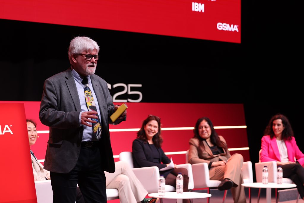 An older man with gray hair and glasses speaks on stage, holding notes, highlighting that post-quantum security is an urgent challenge facing the mobile industry. Behind him, four women sit on chairs, smiling in business attire against a red "MWC23" backdrop. Water bottles decorate tables at this professional conference.
