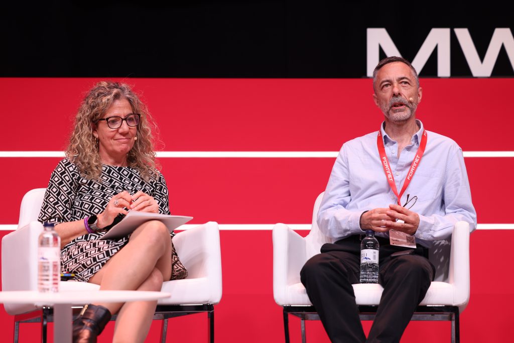 Two people are seated on stage against a red background. The woman on the left, in a patterned dress, holds a notepad, while the man on the right, in a light blue shirt with lanyard, clasps his hands. As bottles of water rest nearby, they discuss how post-quantum security is an urgent challenge for the mobile industry.