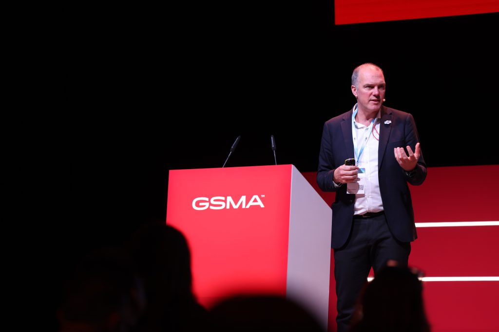 A man in a suit stands on stage beside a red podium labeled "GSMA," gesturing with his left hand and holding a smartphone in his right. The red and black backdrop sets the stage for discussing urgent challenges like post-quantum security facing the mobile industry at this formal conference.