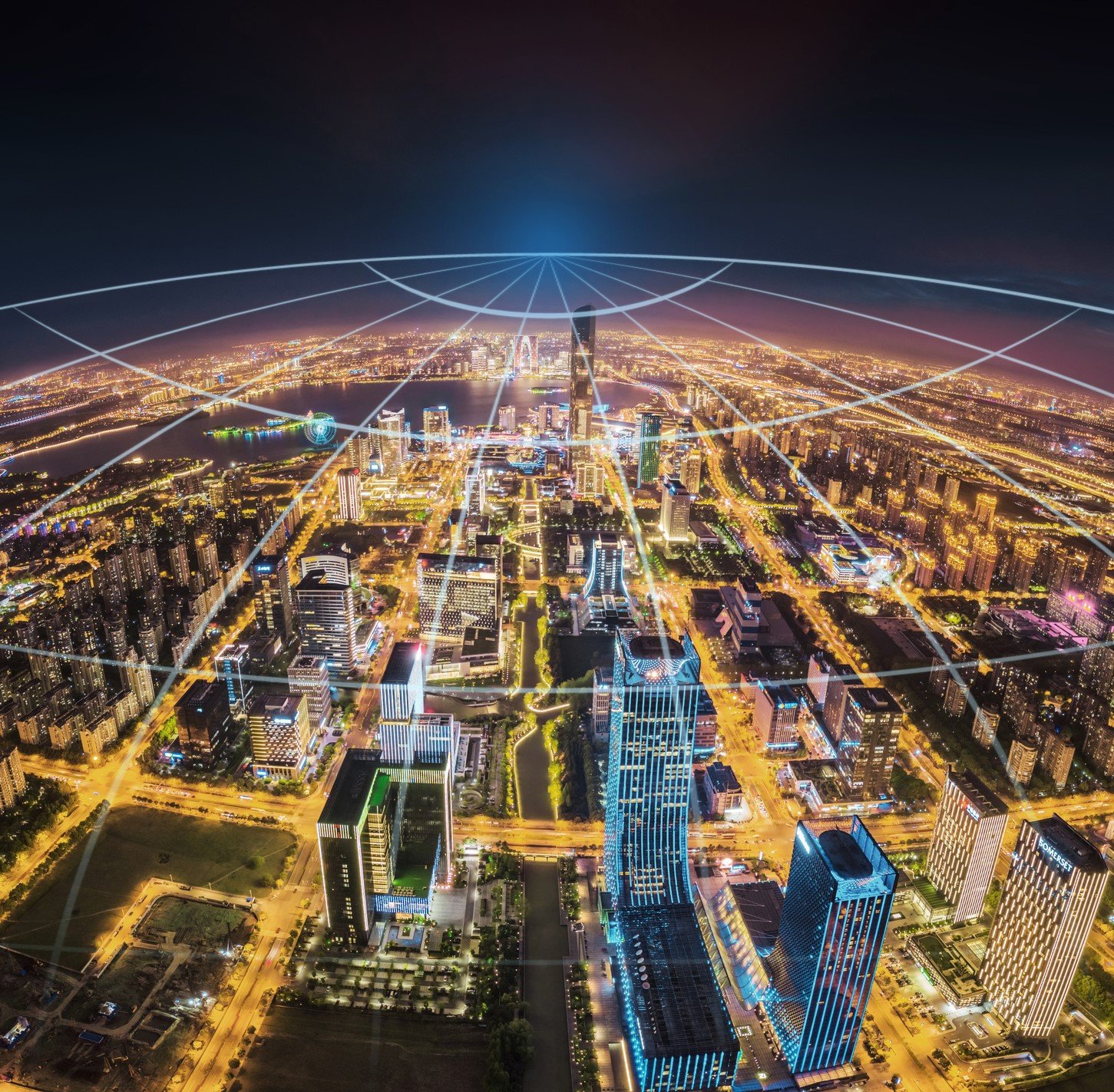 A vibrant cityscape at night viewed from above, with tall buildings illuminated by streetlights. White digital network lines arc over the city, converging at a bright point on the horizon, symbolizing global connectivity and communication.