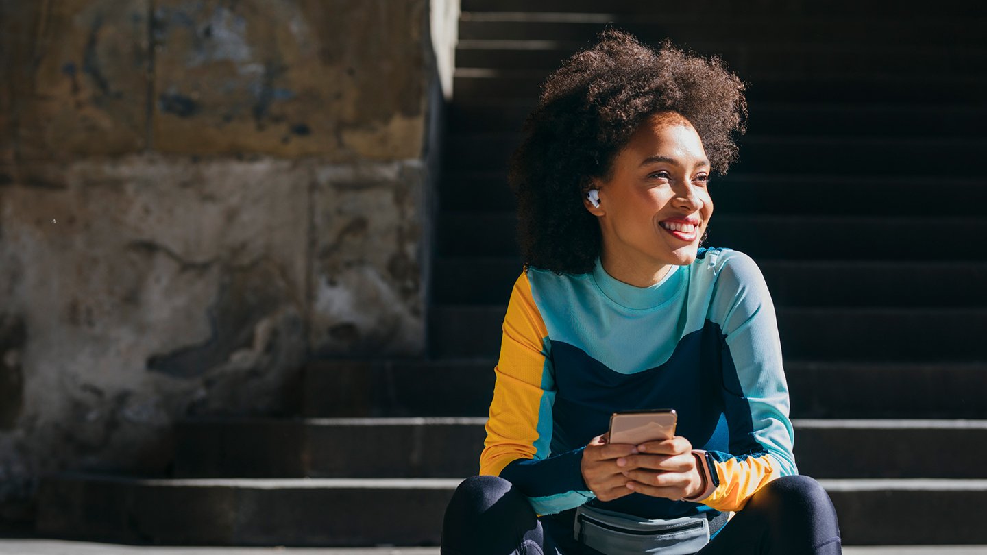 A smiling woman with curly hair, wearing a colorful long-sleeve athletic top and wireless earbuds, sits on outdoor steps, holding a smartphone in both hands. Sunlight highlights her face and hair, and she looks to her right.