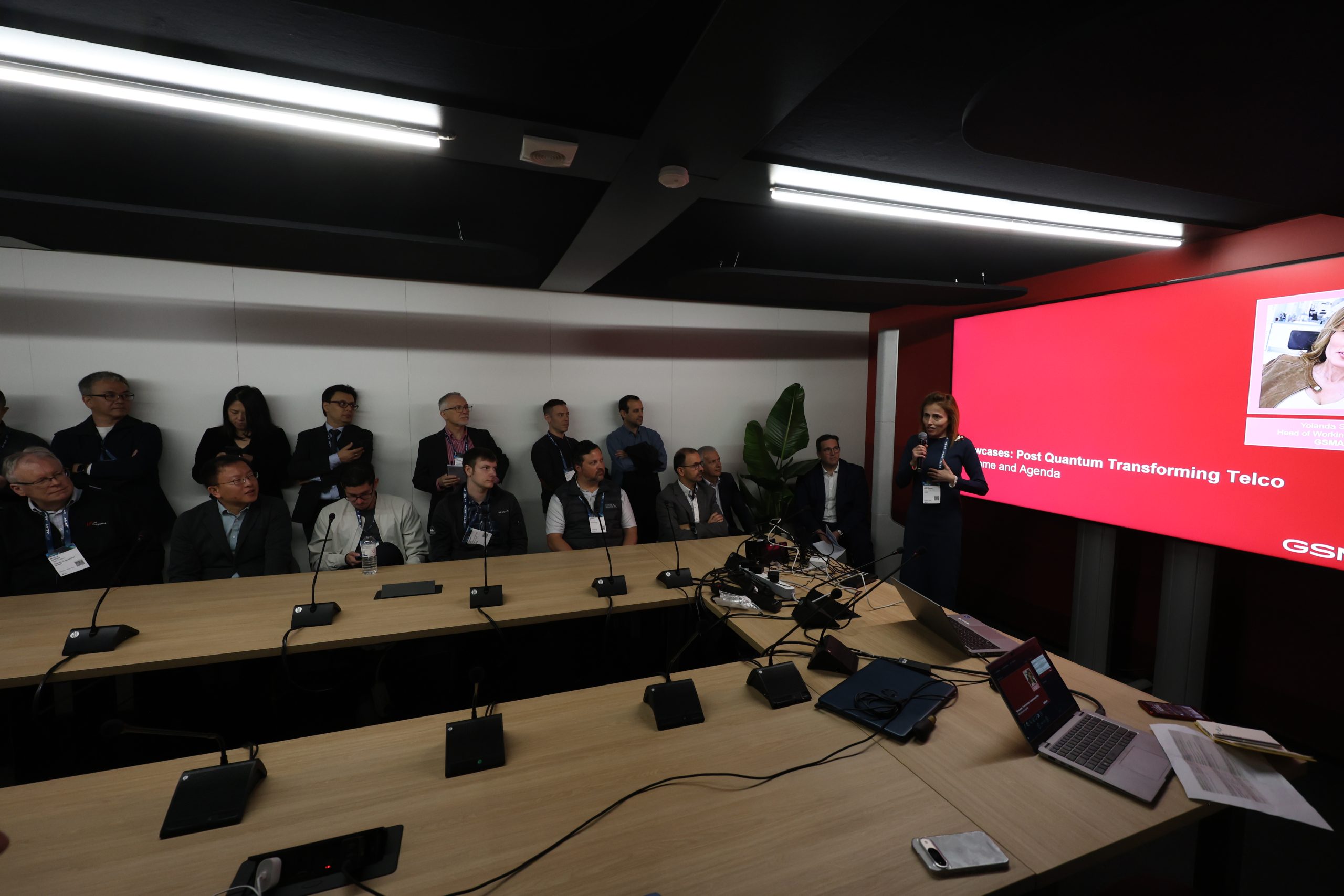 A woman stands presenting beside a large red screen to an audience of business professionals seated and standing around a conference table. The screen highlights “Post-Quantum Transforming Telco,” with tech equipment and laptops visible on the table.