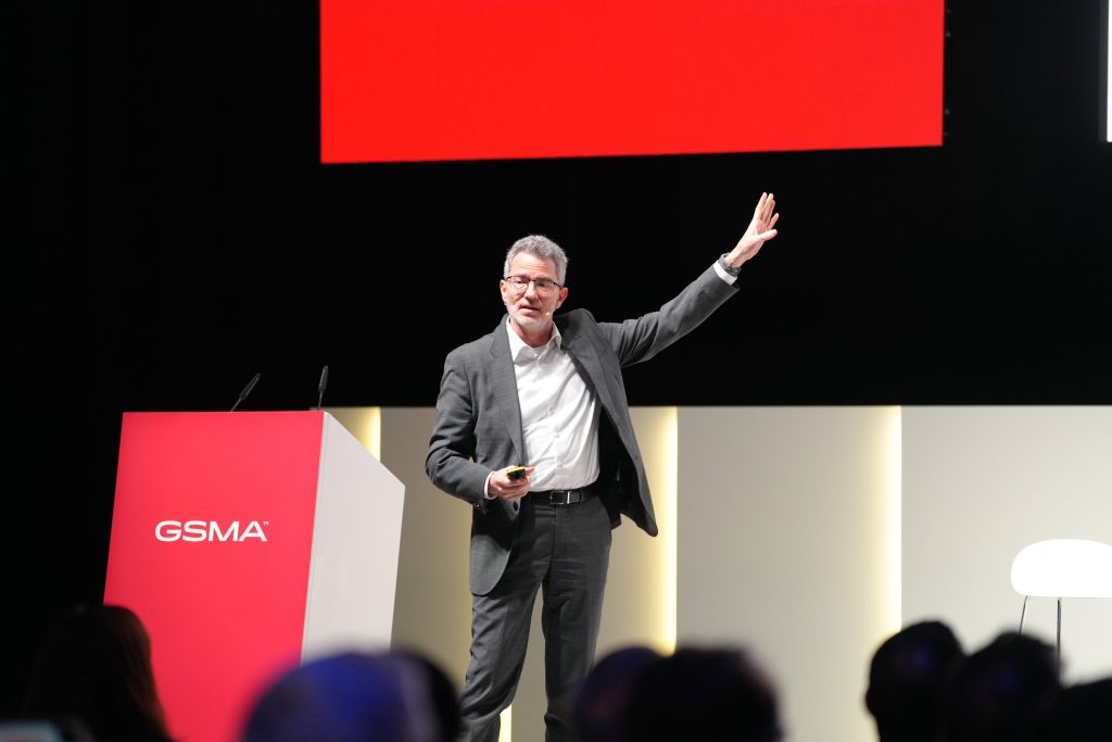 A man in a suit gestures while speaking on stage at a GSMA event, addressing post-quantum cryptography migration as he stands next to a red lectern.