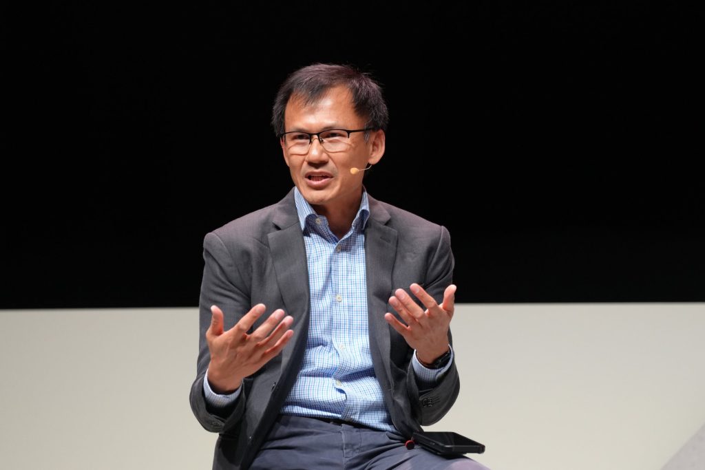 A man in a suit speaks whilst gesturing with his hands, wearing a headset microphone on stage, discussing post-quantum cryptography migration.