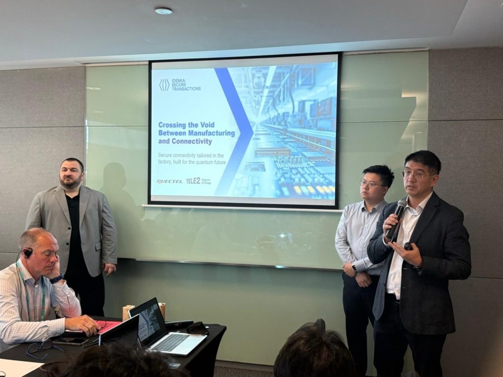 Four men are in a conference room. One is speaking into a microphone while two stand behind him, and another is seated using a laptop with headphones. A presentation slide on a screen reads “Crossing the Void Between Manufacturing and Connectivity”.
