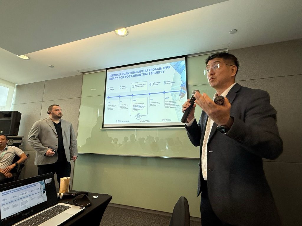 Two men in suits present in a conference room. One speaks into a microphone while gesturing; the other stands near a projected slide detailing a quantum-safe security approach. Attendees and laptops are visible in the foreground.