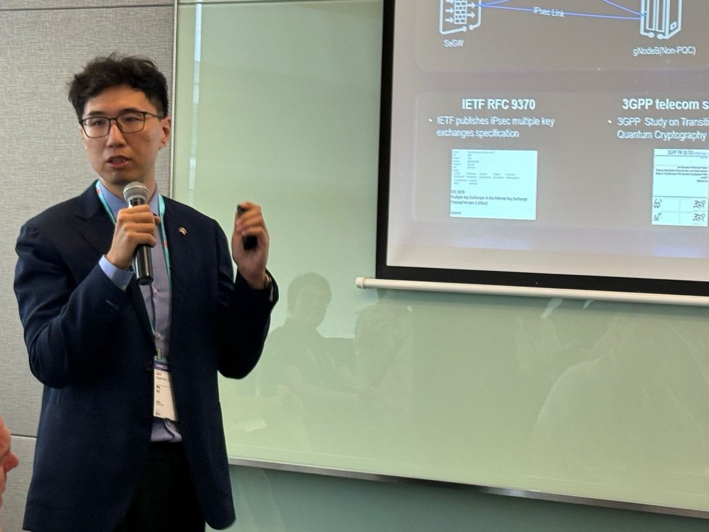 A man in a suit speaks into a microphone and gestures with his hand whilst standing next to a glass wall. Behind him is a projected presentation slide with text and diagrams about telecoms and cryptography standards. Seated audience members are faintly visible in the reflection.