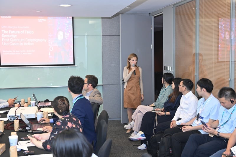 A woman stands and speaks into a microphone in a modern conference room, addressing seated attendees. A presentation slide titled "The Future of Telco Security" is projected on the wall. Attendees listen, take notes, and look towards the presenter.
