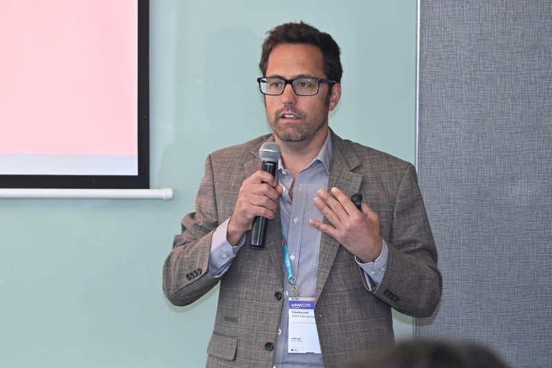 A man in a grey blazer and glasses speaks into a microphone at a conference, gesturing with his left hand. He wears a name badge and stands in front of a projection screen and a blue-grey wall panel.