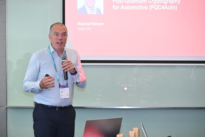 A man in a light blue shirt stands holding a microphone and a presentation remote, speaking in front of a screen displaying a presentation titled “Post-Quantum Cryptography for Automotive (PQC4Auto)” by Maxime Flament, SGAA CTO.