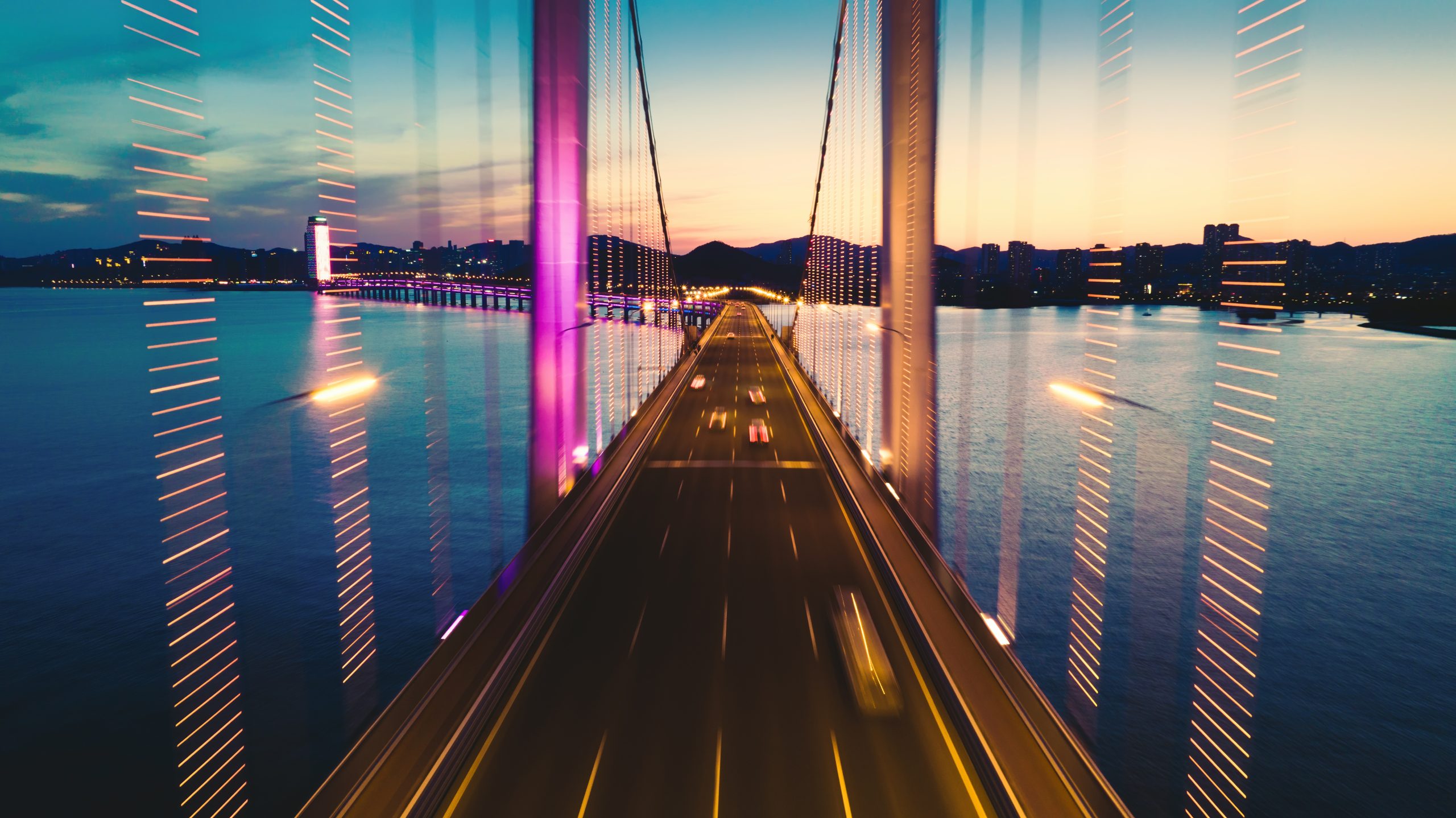A long suspension bridge stretches over dark water at sunset, its structure secured like post-quantum cryptography against the night. Motion-blurred streaks of light from moving cars pass the towers, which are illuminated with bright purple and white lights against the evening sky.