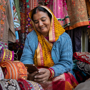 A woman in a yellow and pink saree with a blue jumper sits among colourful fabrics, smiling whilst looking at a mobile phone. She has a red bindi on her forehead and is surrounded by vibrant textiles at a market stall.